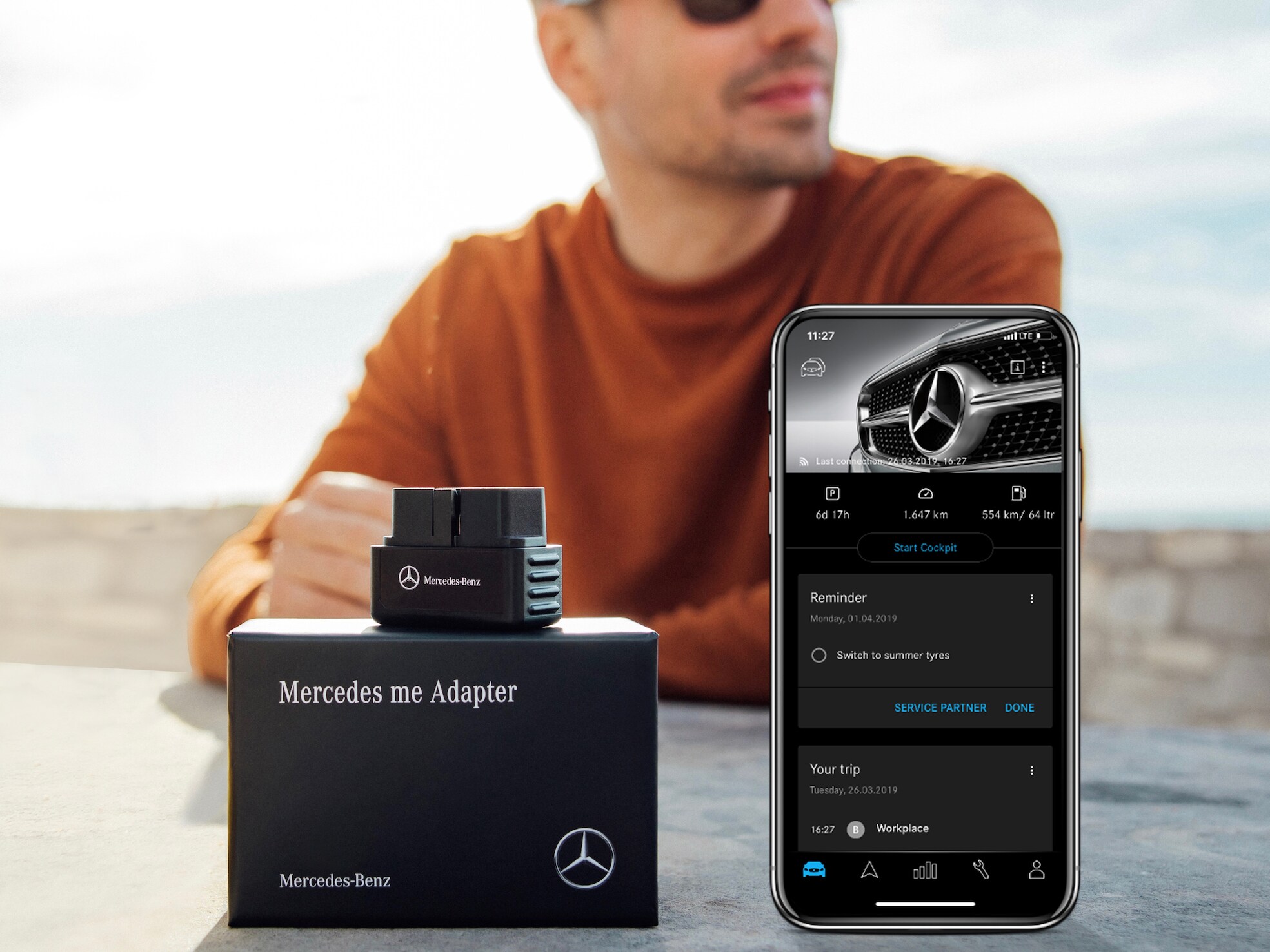 선글라스를 쓴 남성이 앉아 있는 테이블 위에 Mercedes-Benz Adapter와 Mercedes-Benz Adapter 앱이 열린 스마트폰이 보입니다.