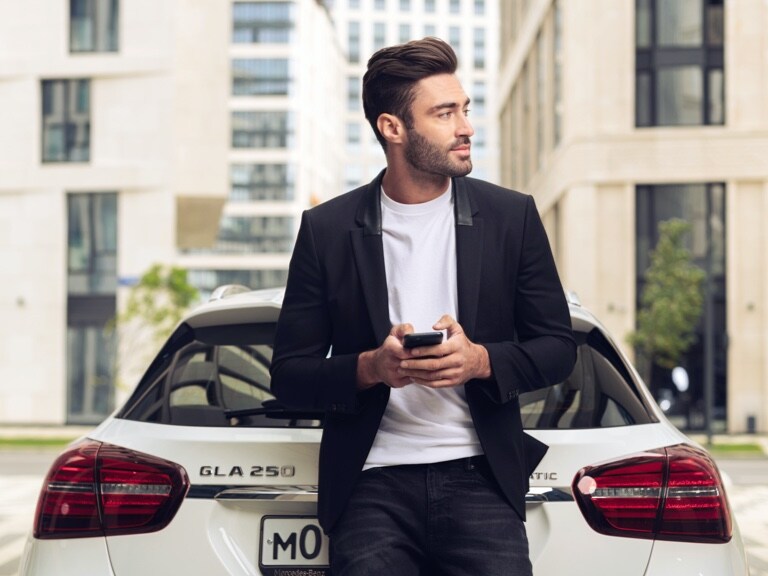 한 남성이 자신의 Mercedes-Benz GLA에 기대어, 스마트폰의 Mercedes me App과 내비게이션 관련 디지털 서비스 상품을 이용하여 목적지의 날씨를 확인하고 있습니다.